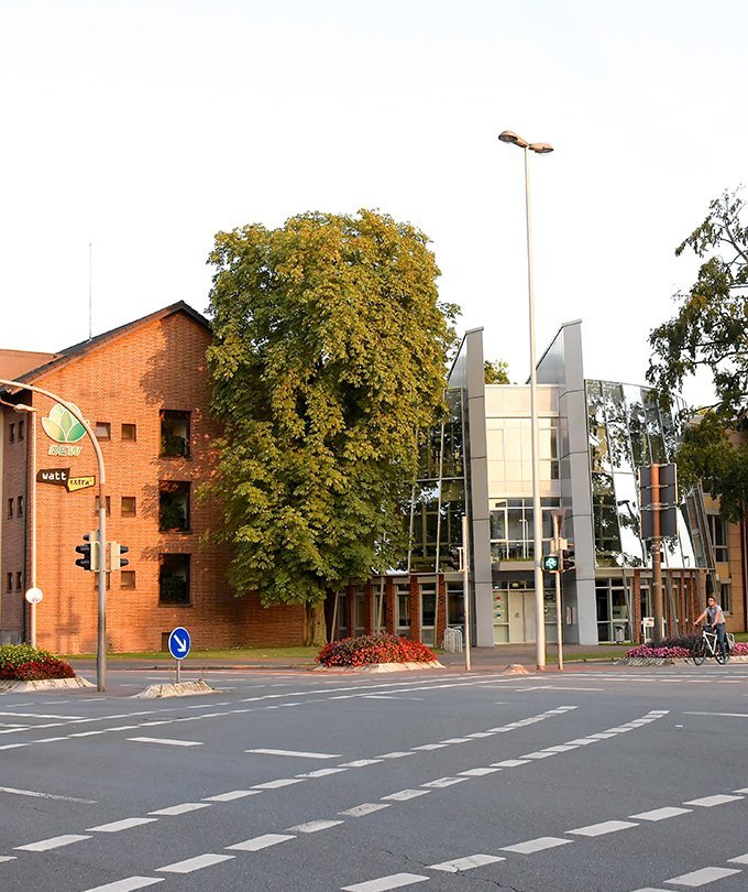 Ein modernes Gebäude mit großen Glasfenstern steht neben einem roten Backsteingebäude, umgeben von Bäumen und Blumenbeeten.