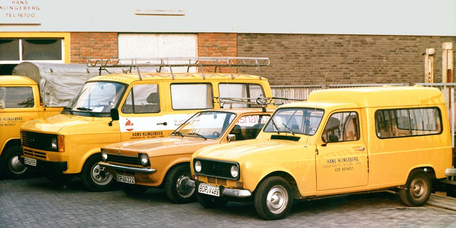 Drei gelbe Fahrzeuge stehen nebeneinander auf einem gepflasterten Parkplatz vor einer Backsteinmauer.