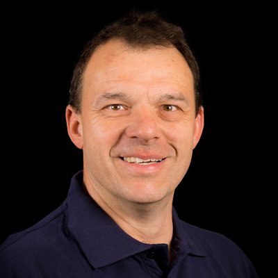 Ein lächelnder Mann mit kurzen, braunen Haaren trägt ein dunkelblaues Polohemd vor einem schwarzen Hintergrund.