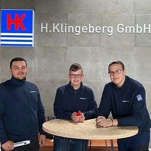 Drei junge Männer stehen an einem runden Tisch in einem modernen Büro, lächeln und tragen dunkelblaue Arbeitsjacken.